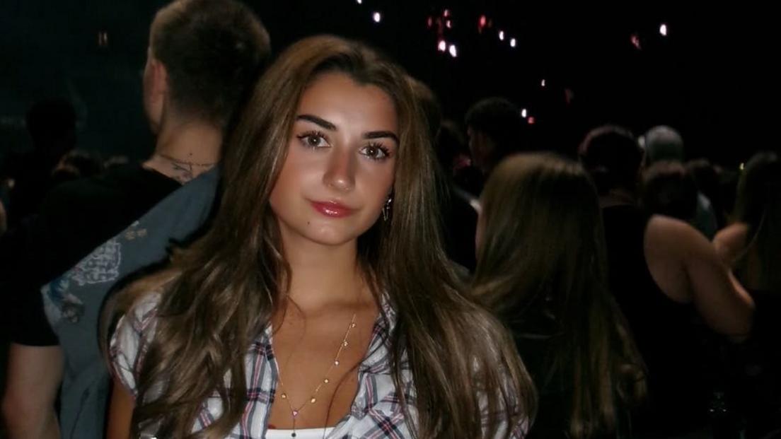Evie Lewis, a young woman standing in what appears to be a music venue. She has long dark hair and is wearing a checked short sleeved top over a white vest with a thin gold necklace. Behind her there are lots of people looking at what might be a stage off picture.