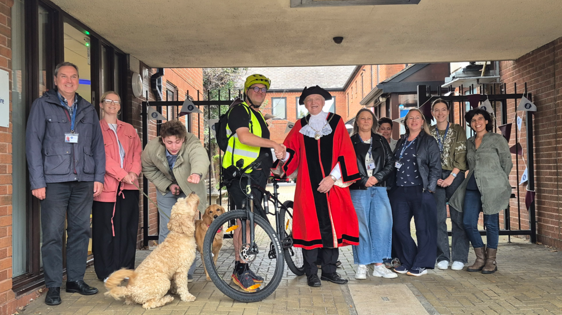 A group of people posing and smiling to camera by a block of houses with iron railings behind. Ed is in cycling gear and a high vis jacket and helmet by his bike and he is shaking hands with a mayor in full regalia. There are two dogs, a golden retriever and a cockapoo also in the pictures