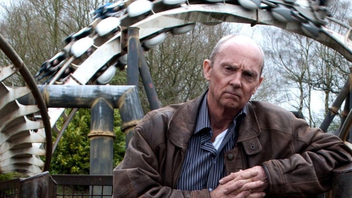 John Wardley, standing in front of a rollercoaster, with his arms crossed in front of him. He has on a brown leather coat, blue and white shirt and white T-shirt underneath. He has short hair. 