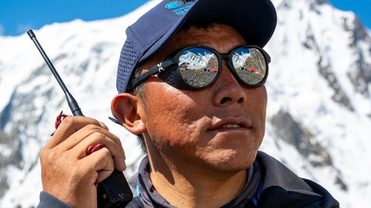 A Nepalese man wearing a cap and sunglasses, holding a walkie-talkie with snowy mountains behind him
