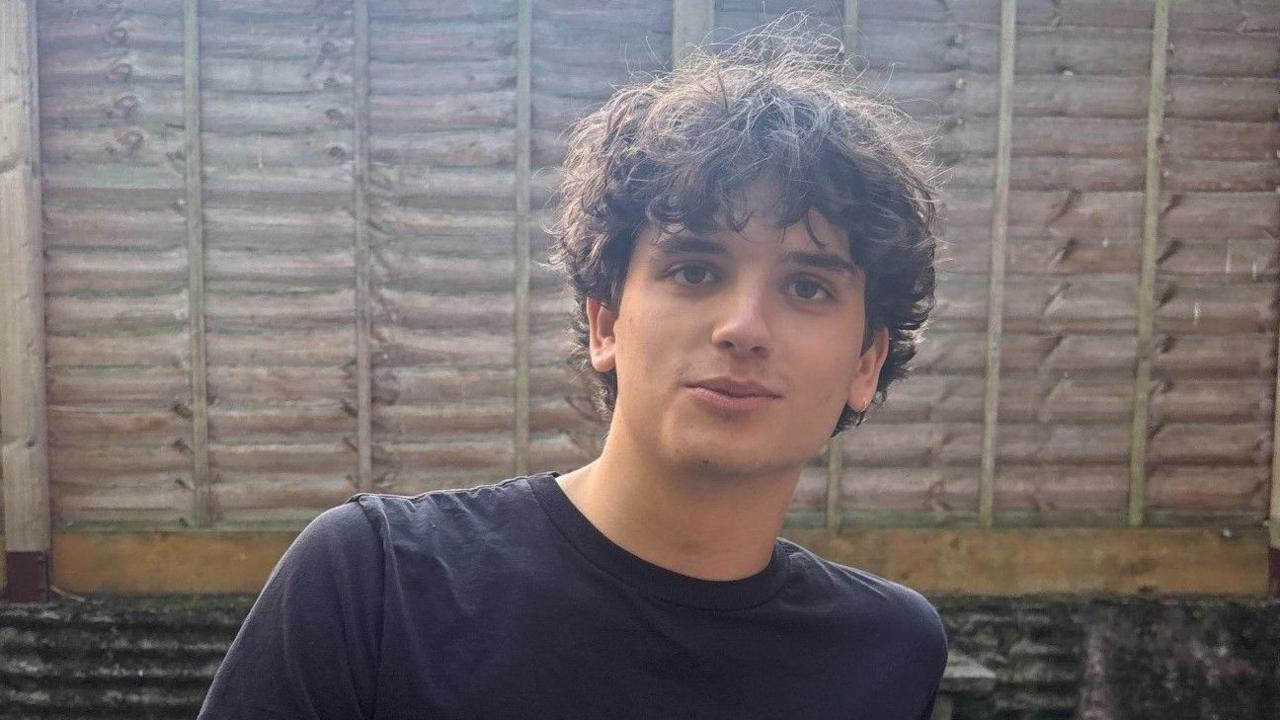 Headshot of Archie Forde, 19. He is sitting on a bench in a garden with a fence behind him. He wears a black T-shirt, has longish dark brown hair, brown eyes and looks straight at the camera.