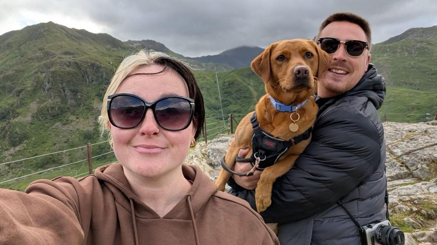 A selfie photograph of Lauren Evans (left) and her boyfriend and dog (right) in the green mountains in north Wales. Lauren has blonde and black hair and smiles at the camera wearing black wide sunglasses. She wears a brown hoodie while her boyfriend wears a black and grey puffer coat and has a camera strapped across his body while he holds their dog in his arms. He has brown short hair and wears black sunglasses. 