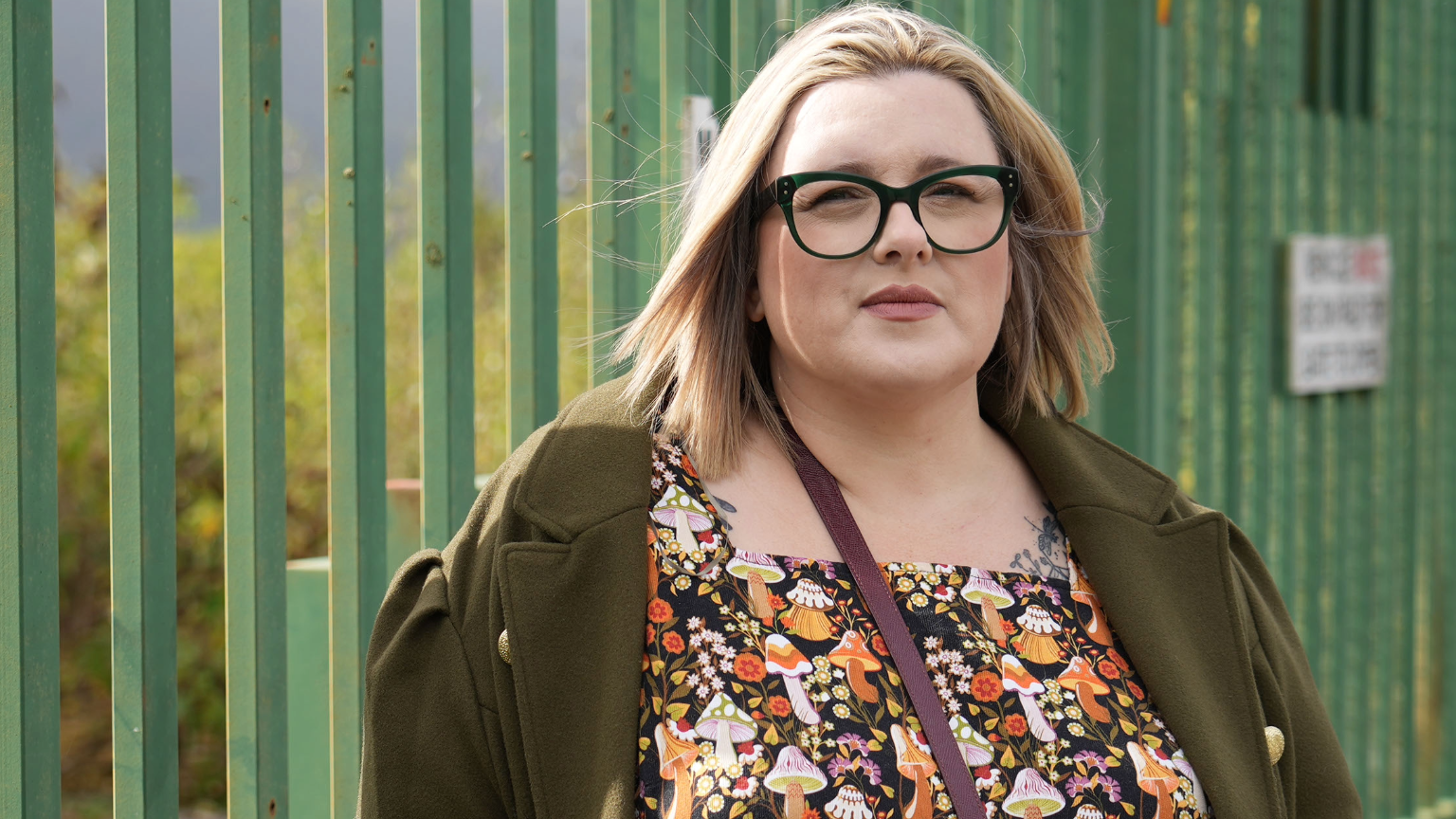 Community councillor Katie Witherden is a white woman, with blonde hair and black glasses standing in front of a green fence wearing a green coat and a pattern dress