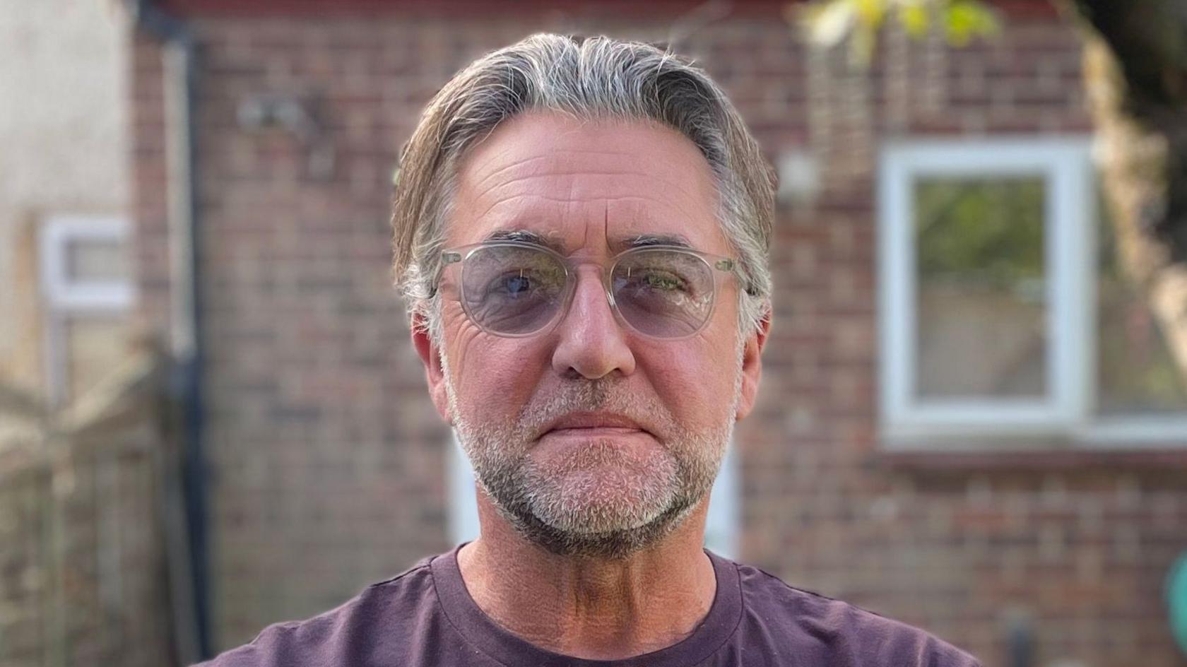 Ritchie Wood has his hair swept backwards and is wearing glasses and a dark purple T-shirt. He is standing in his back garden with his brick house behind him.