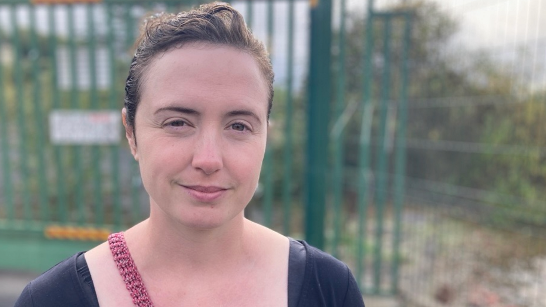 Natalie Dugdale is a white lady with brown hair wearing a blue t-shirt and a red handbag in front of a wooded area