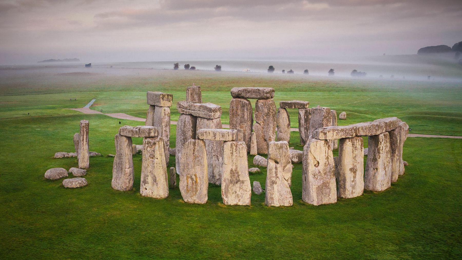 The image shows Stonehenge. There is a misty sky behind with the full circles of the ancient monument in the foreground. Four sets of two large upright stones, each with another large stone resting across the top, which make up part of the outer circle. The even larger stones of the inner area are visible above those of the outer circle. Green grass surrounds the monument on all sides.