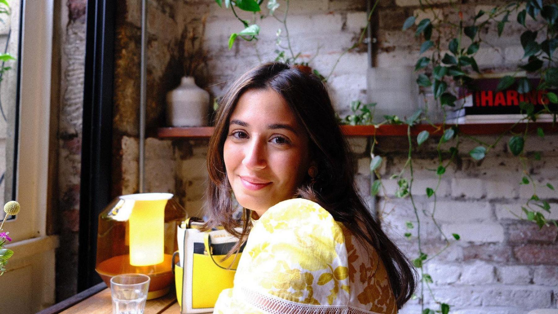 Girl with brunette hair and a yellow top smiles inside a dimly lit coffee shop.