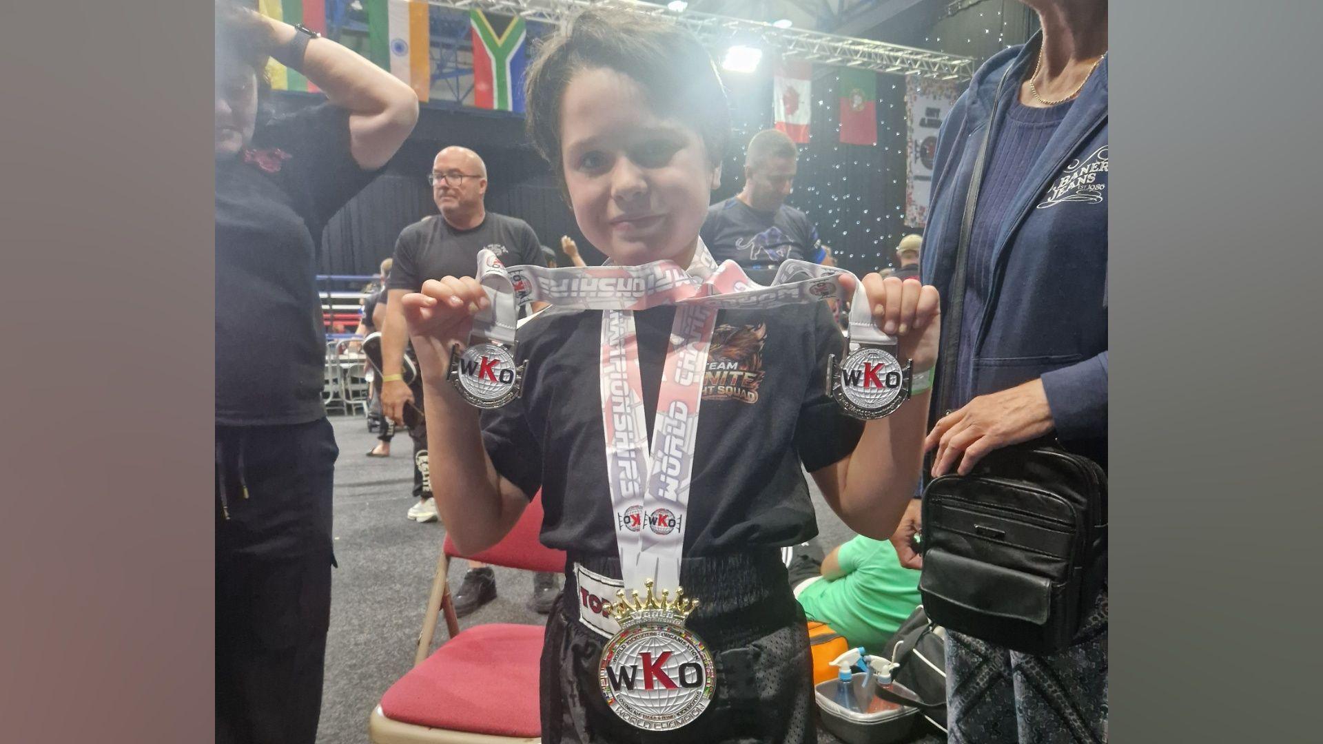 A 10-year-old girl holds up two smaller medals, which are around her neck, and smiles at a kickboxing competition. She has a larger medal, which is hanging from her neck. She is wearing a black kickboxing outfit and coaches and other competitors can be seen behind her. She has short brown hair.