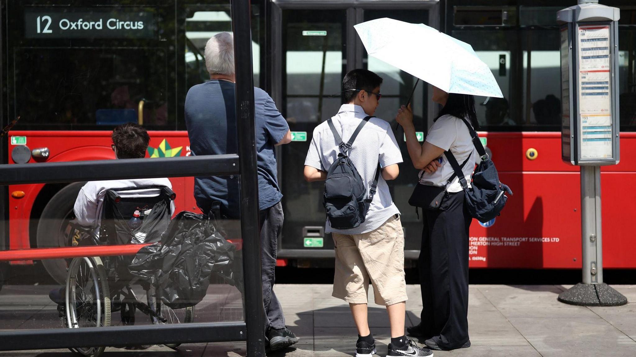 A picture showing people at a bus stop in London with the number 12 bus to Oxford Circus at the stand. From left to right there is a man in a white shirt who is a wheelchair user, a man in a blue shirt and black trousers leaning on the cover and two people under a white umbrella - one is a man in a light coloured shirt and brown trousers, and a woman in a white shirt and black trousers also wearing a backpack.