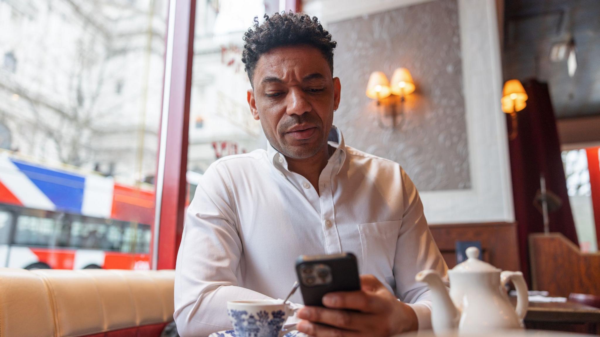 A middle-aged man is having a coffee at a café and sitting next to a window. He is holding his phone and checking something carefully on it