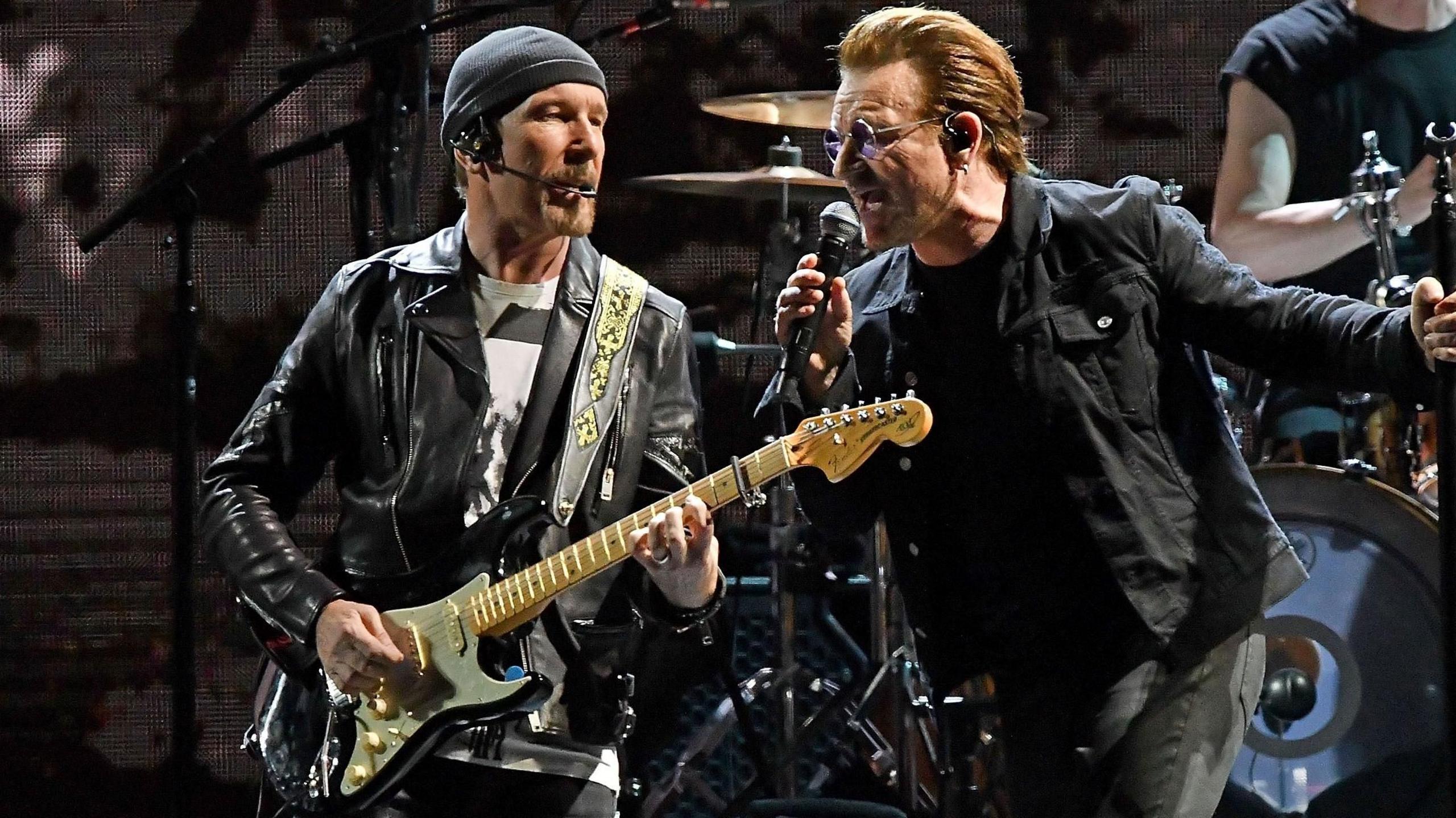 The Edge in black leather jacket and grey hat, playing a guitar, alongside Bono singing into microphone.