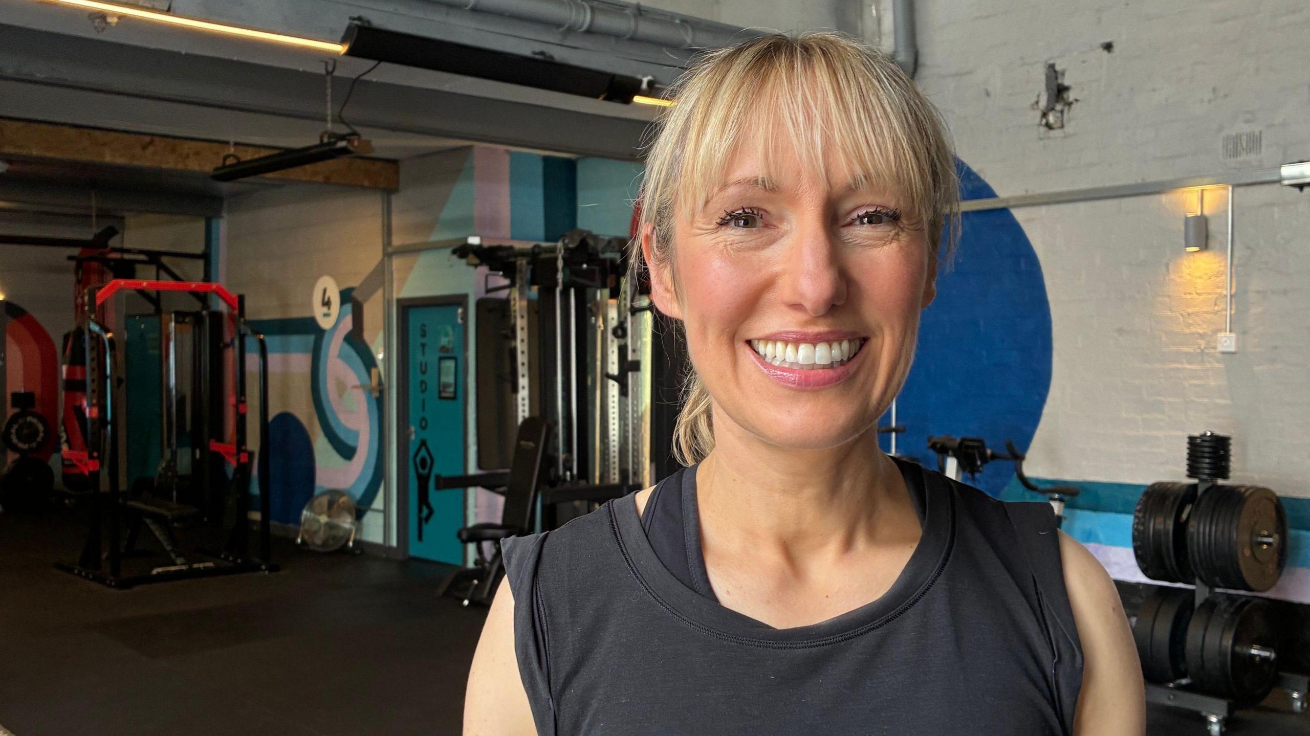 Bev Wilson is in a gym, wearing workout clothes. She is smiling at the camera and has blonde hair in a ponytail. 
