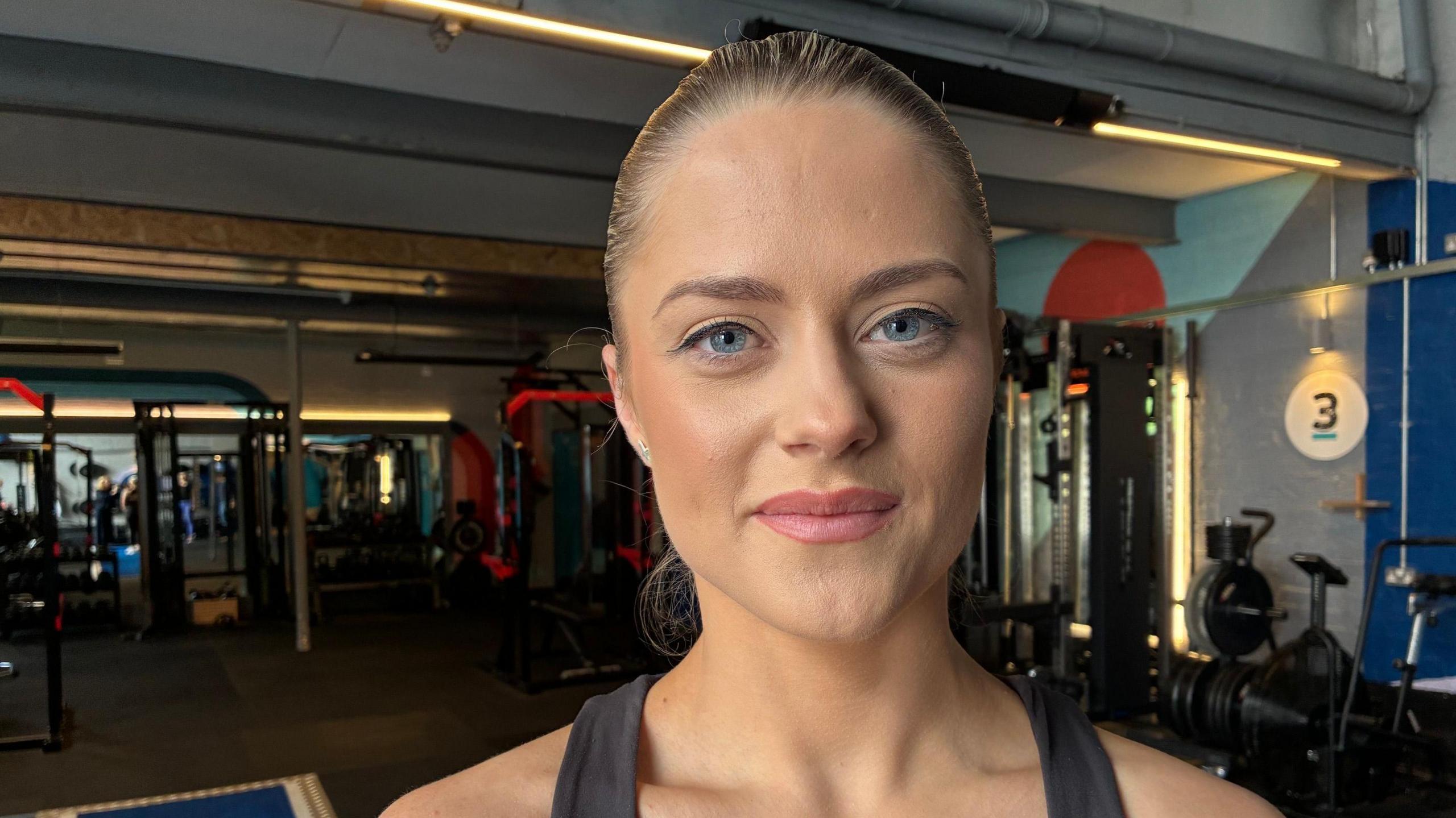 A dark haired woman with her hair sleeked back behind her head wears workout clothes and stands in a gym. She is looking directly at the camera and smiling. 
