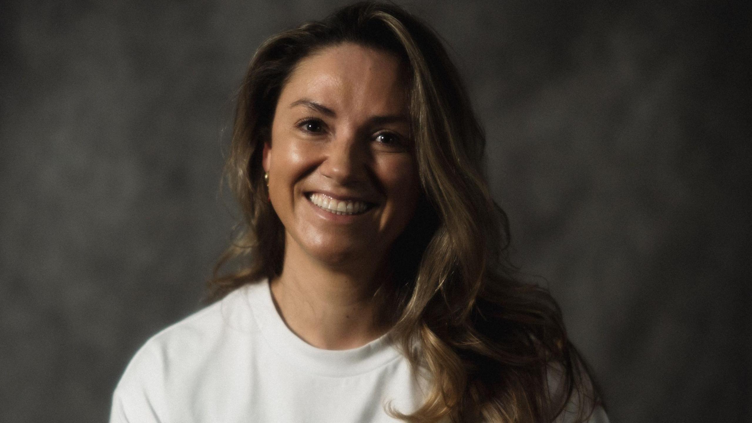 Picture of a woman smiling in a white t-shirt. She has brown hair and is in front of a grey backdrop.