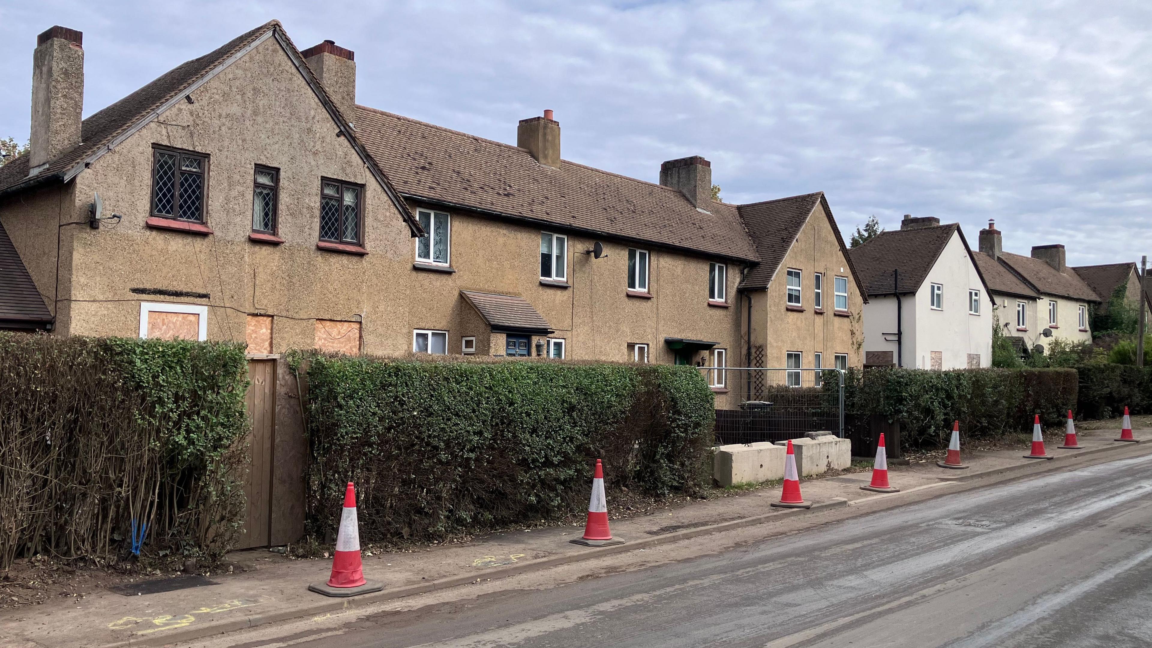 Terraced houses in Manor Road, many of which appear empty and boarded up. There is a line of traffic cones on the pavement in front of them.