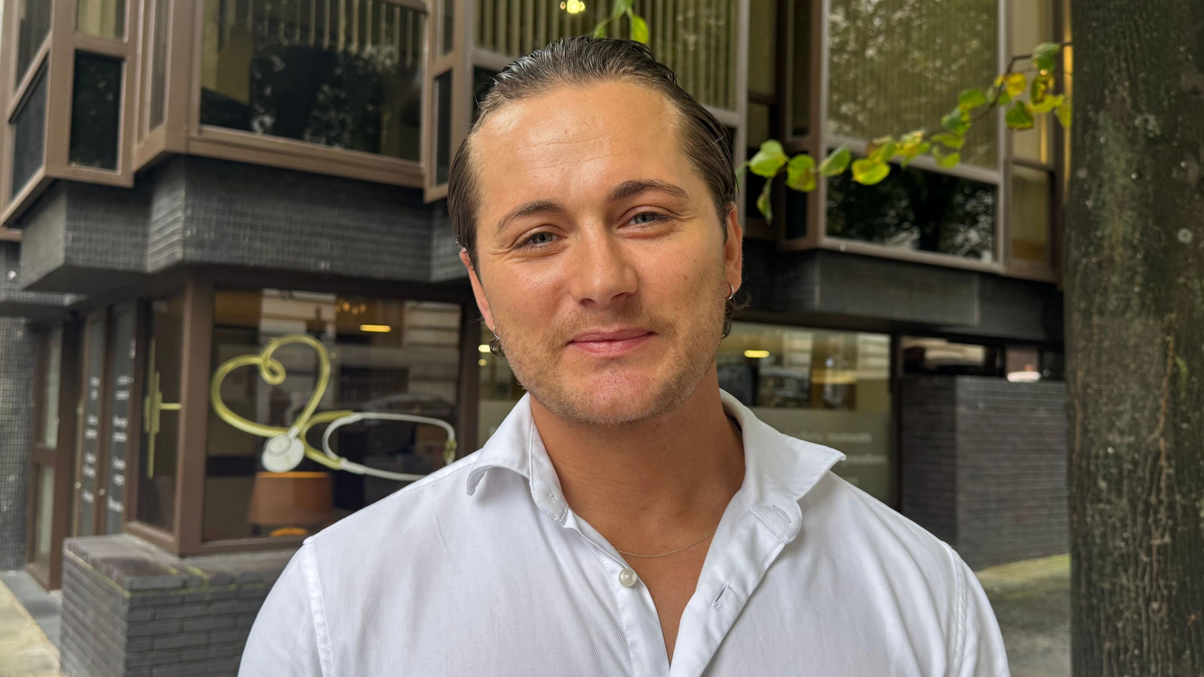 Max Longman, a man in his thirties, with his hair slicked back, wearing a white shirt opened at the collar, standing in front of a building