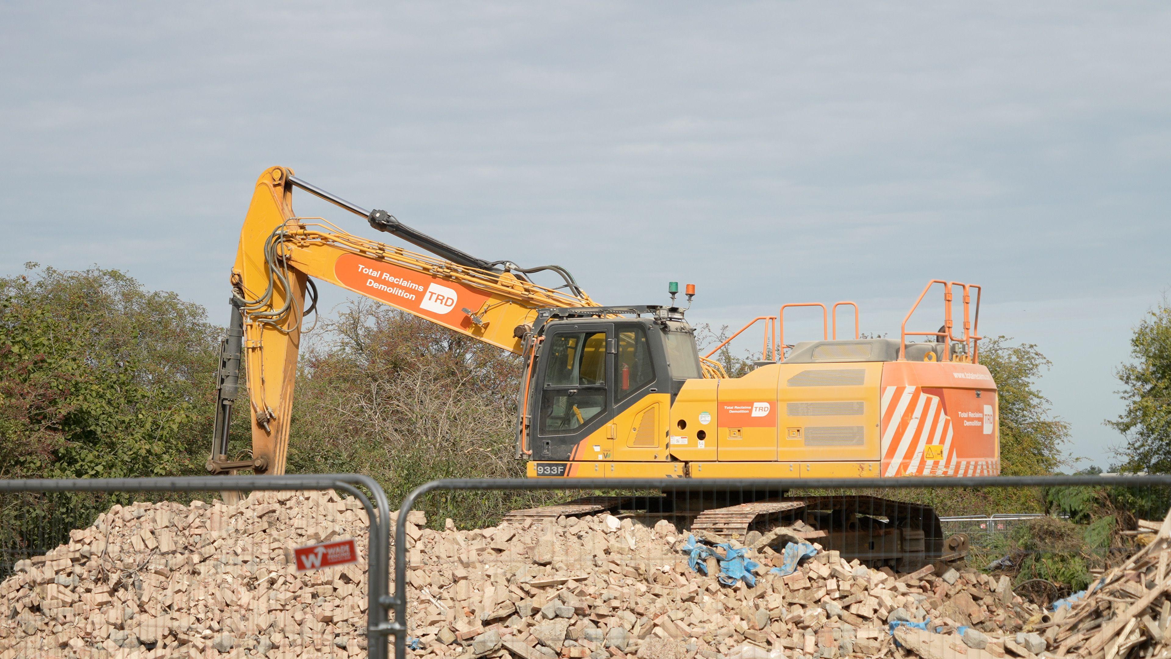 A large yellow digger perched on top of a big pile of rubble. It is cloudy above and there are trees in the background.