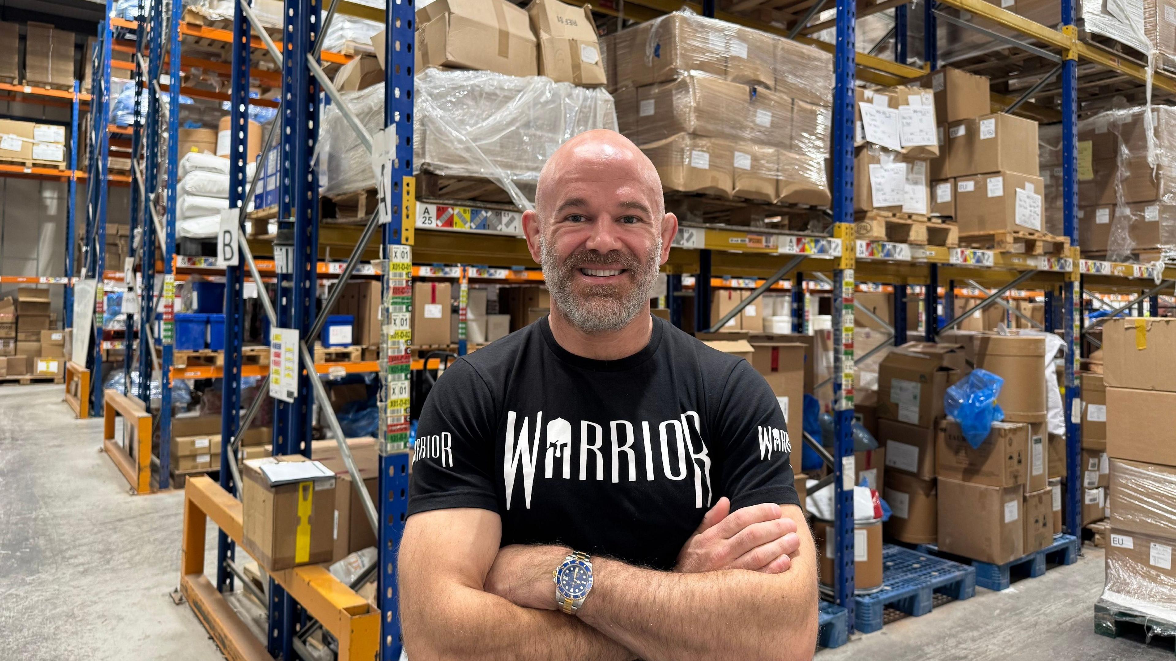 Kieran Fisher, a bald man with a short grey beard wearing a black T-shirt with the word WARRIOR on the front, standing smiling in a warehouse. Behind him are cardboard boxes stacked on blue metal shelves.
