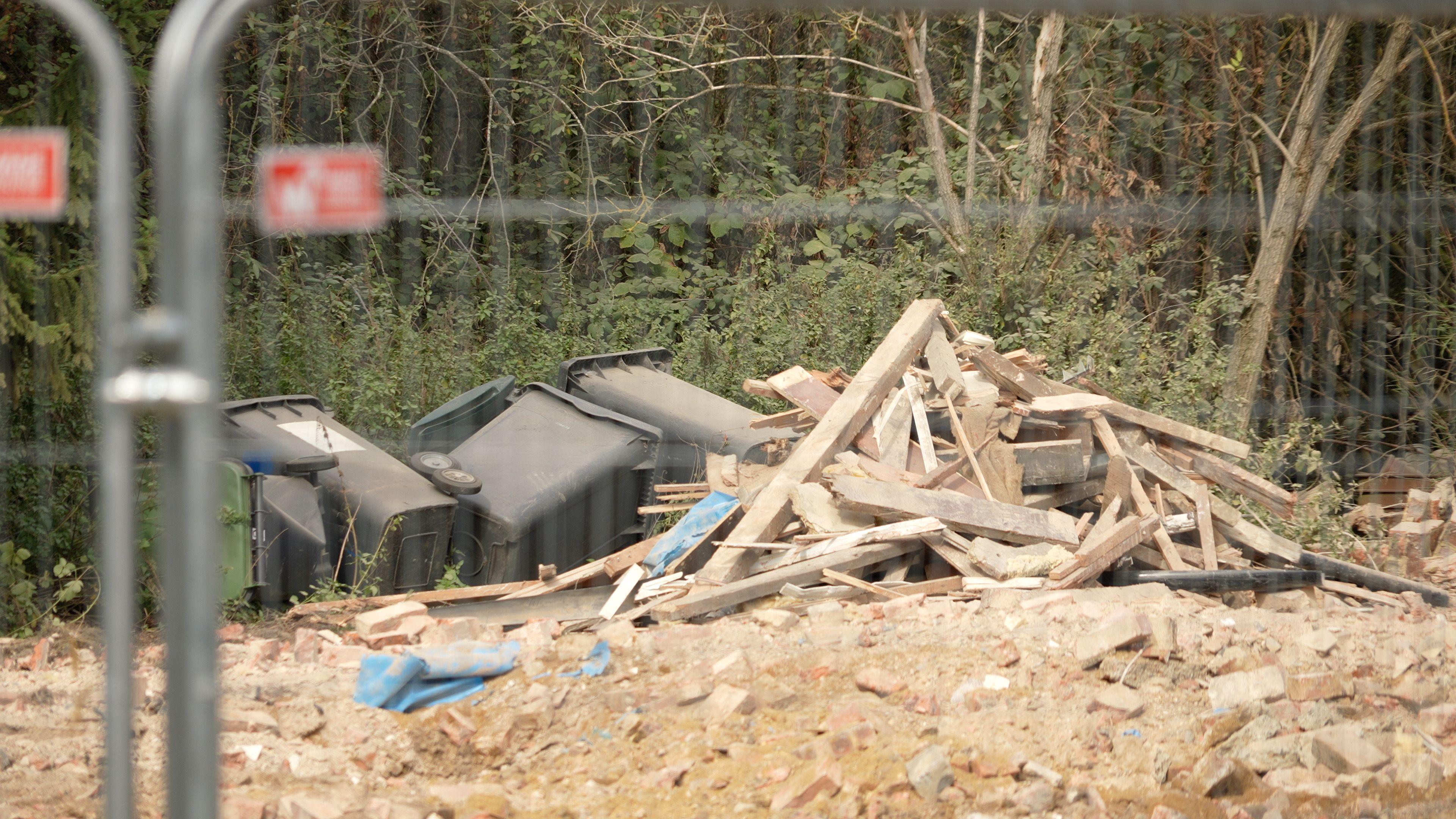 A pile of rubble and wood from where a house has been demolished, pictured behind a metal fence. Several black bins have been toppled onto their sides.