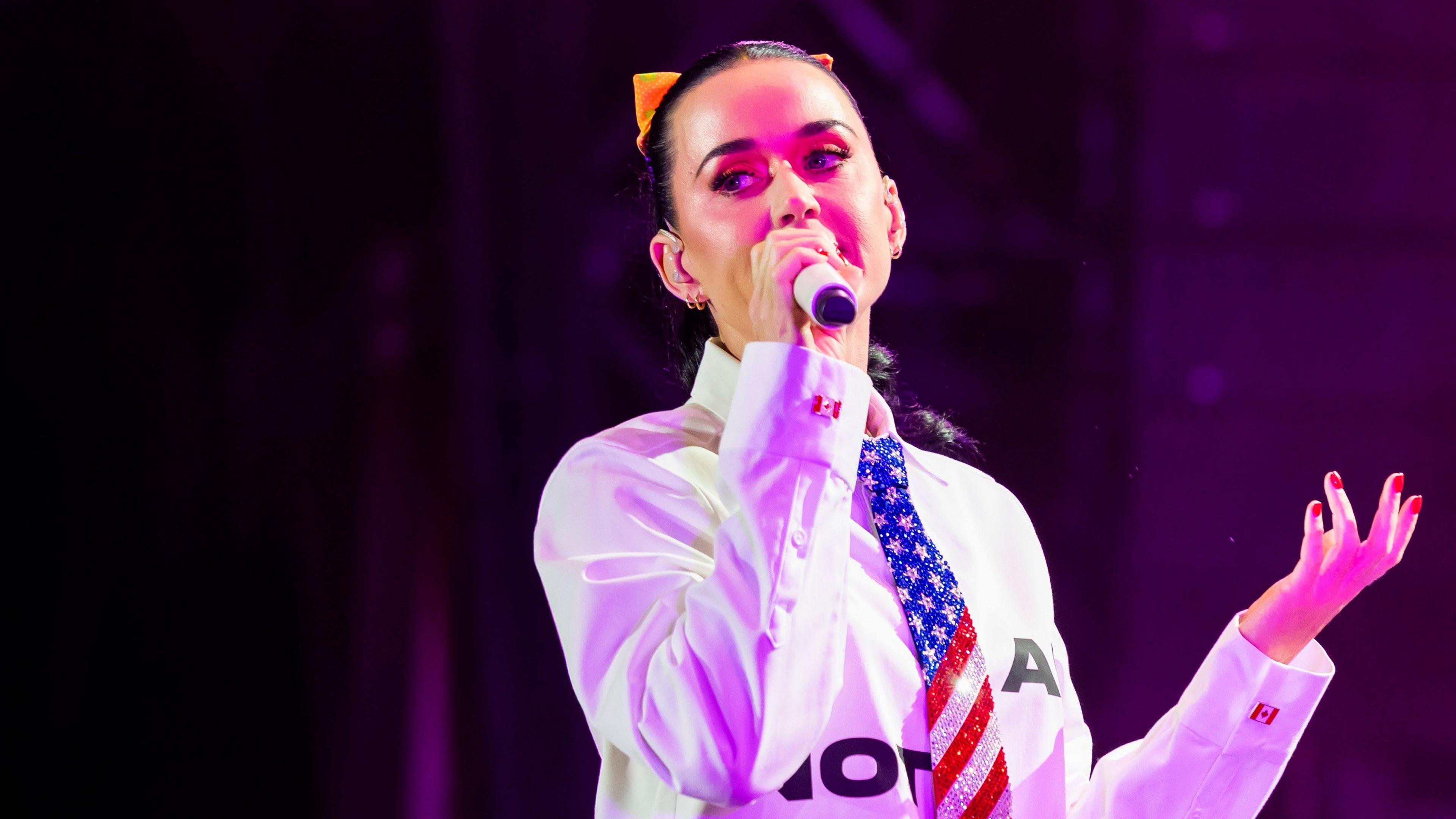 Katy Perry sings on stage. She is wearing a white shirt with an American patterned tie. 