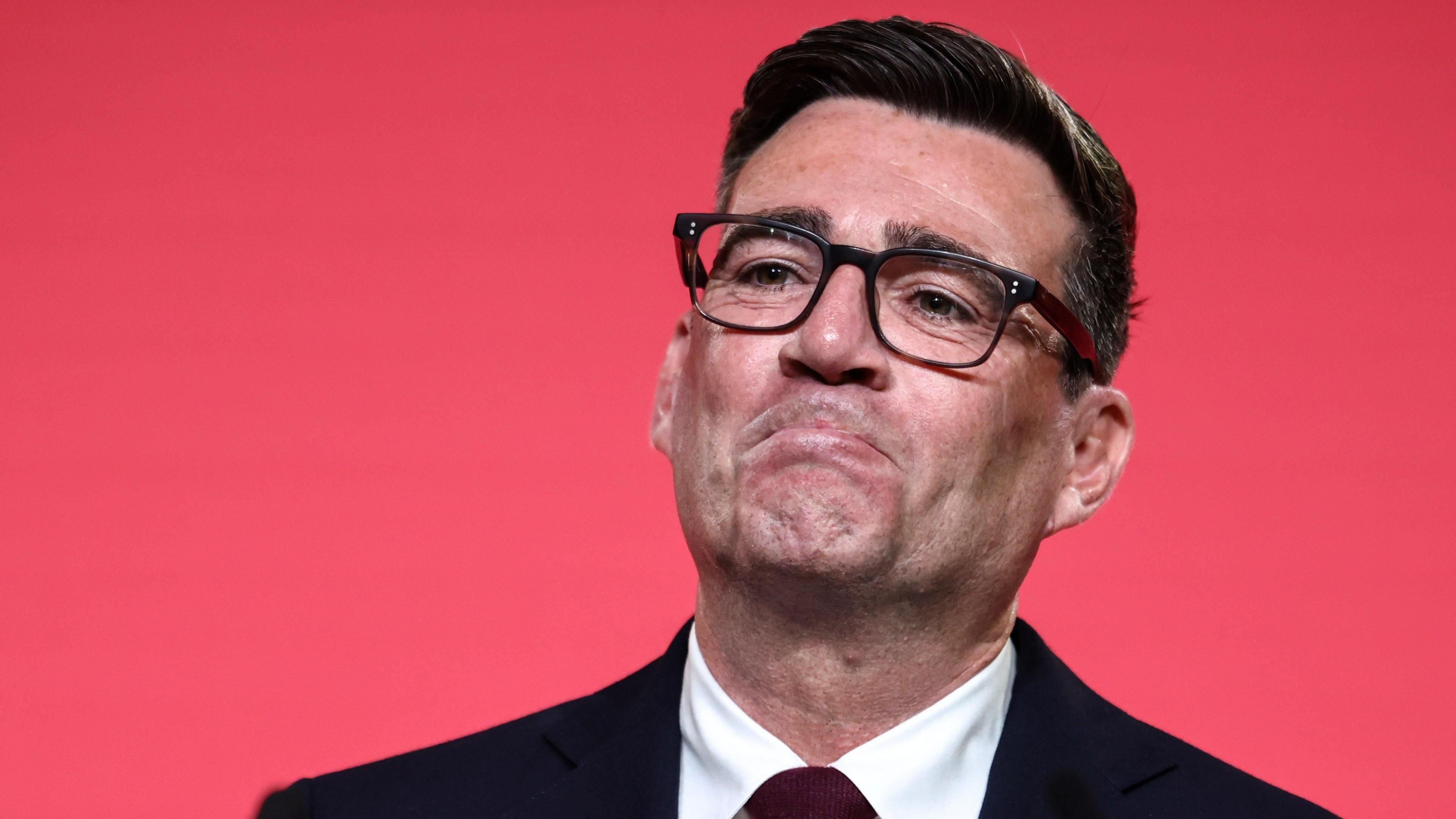 Andy Burnham looking pleased as he reacts to becoming Labour leader. He is wearing black rimmed glasses and a dark suit and tie, against a red background.