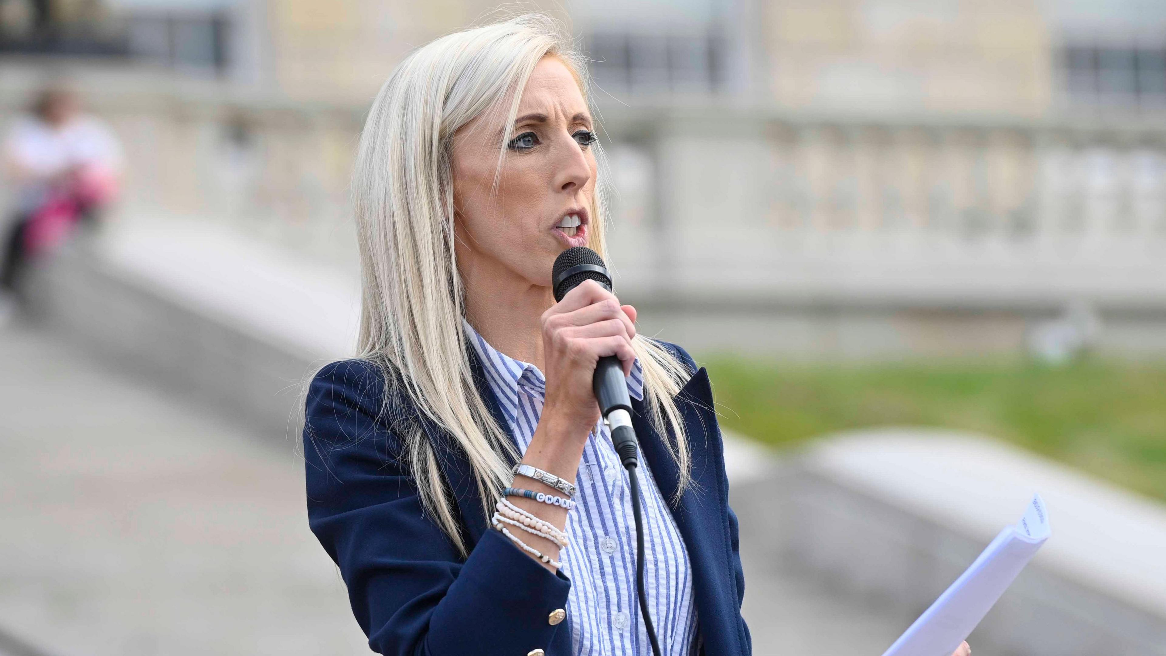 Carla Lockhart addreses an audience with a microphone in one hand, and a sheet of paper in the other. She is wearing a navy blazer, with a blue and white striped shirt
