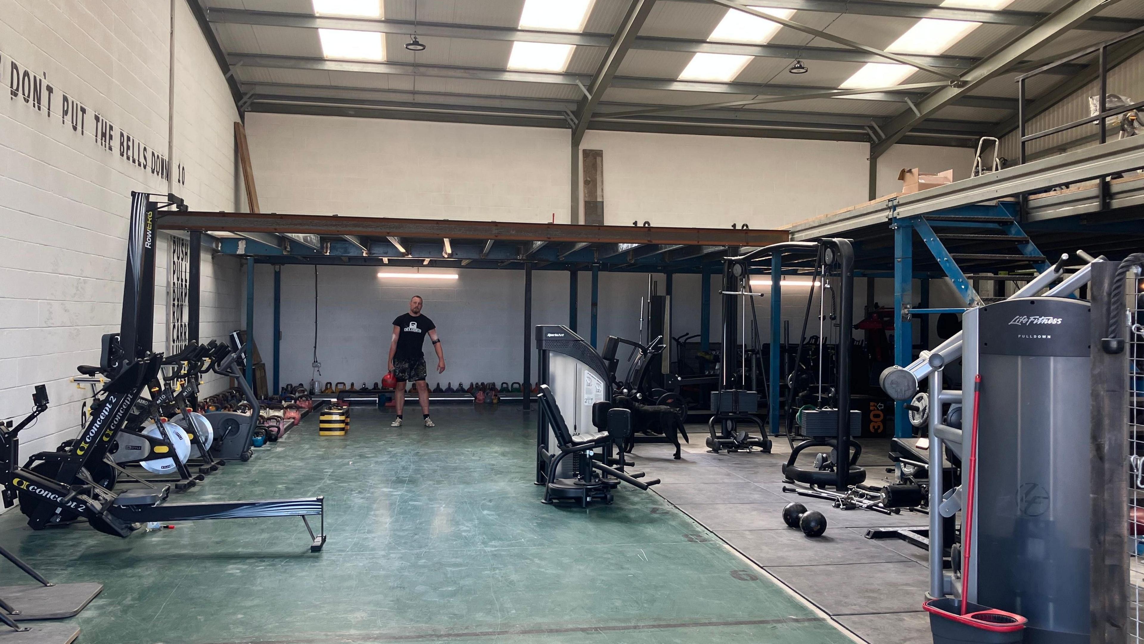 A man is stood under a tall metal frame holding a weight. He is inside a large warehouse type building with exercise equipment around the space