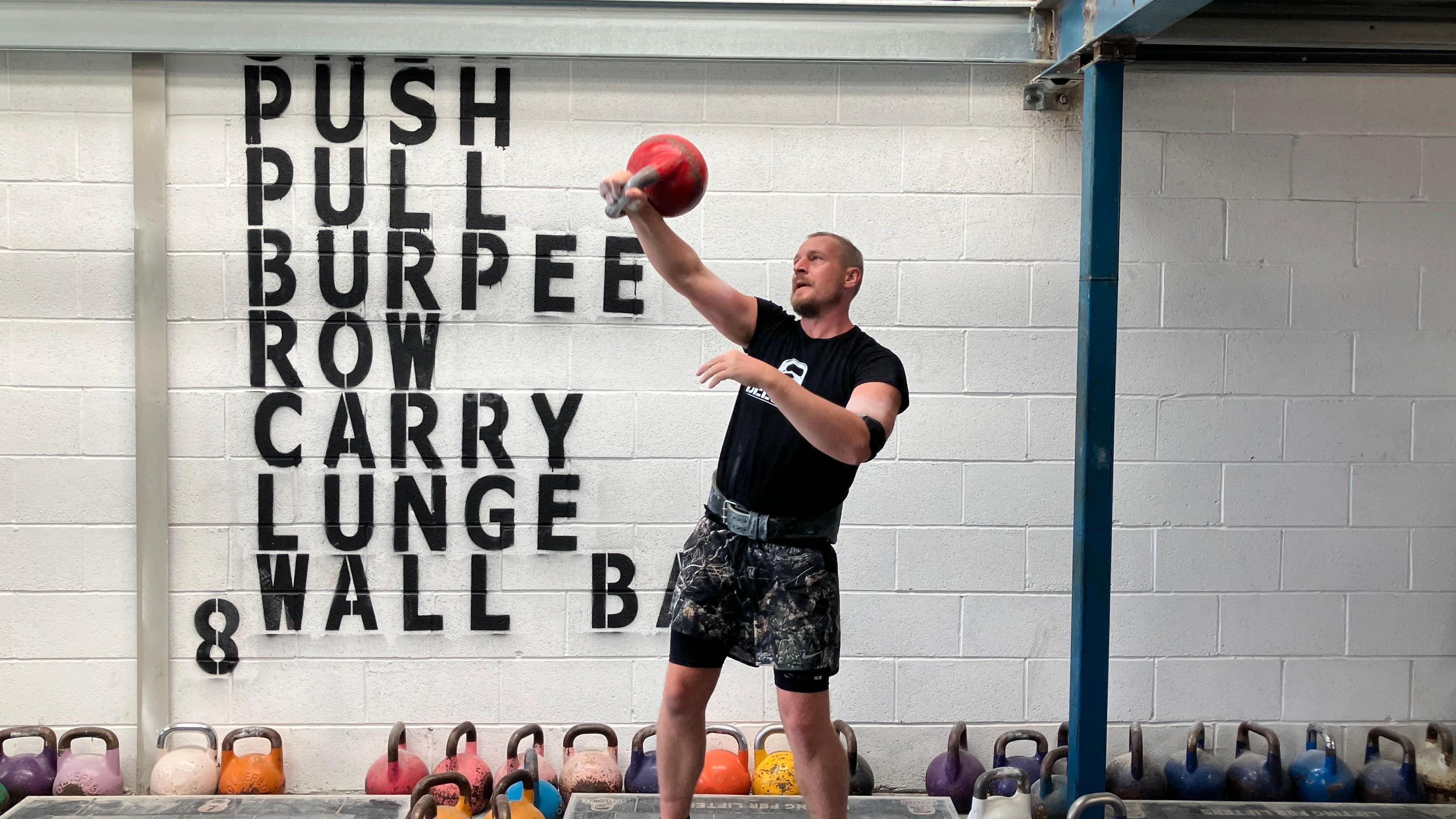 A man holds a red kettleball outstretched in front of him. He is alongside a white wall with the words push, pull, burpee, row, carry, lunge, wall on it