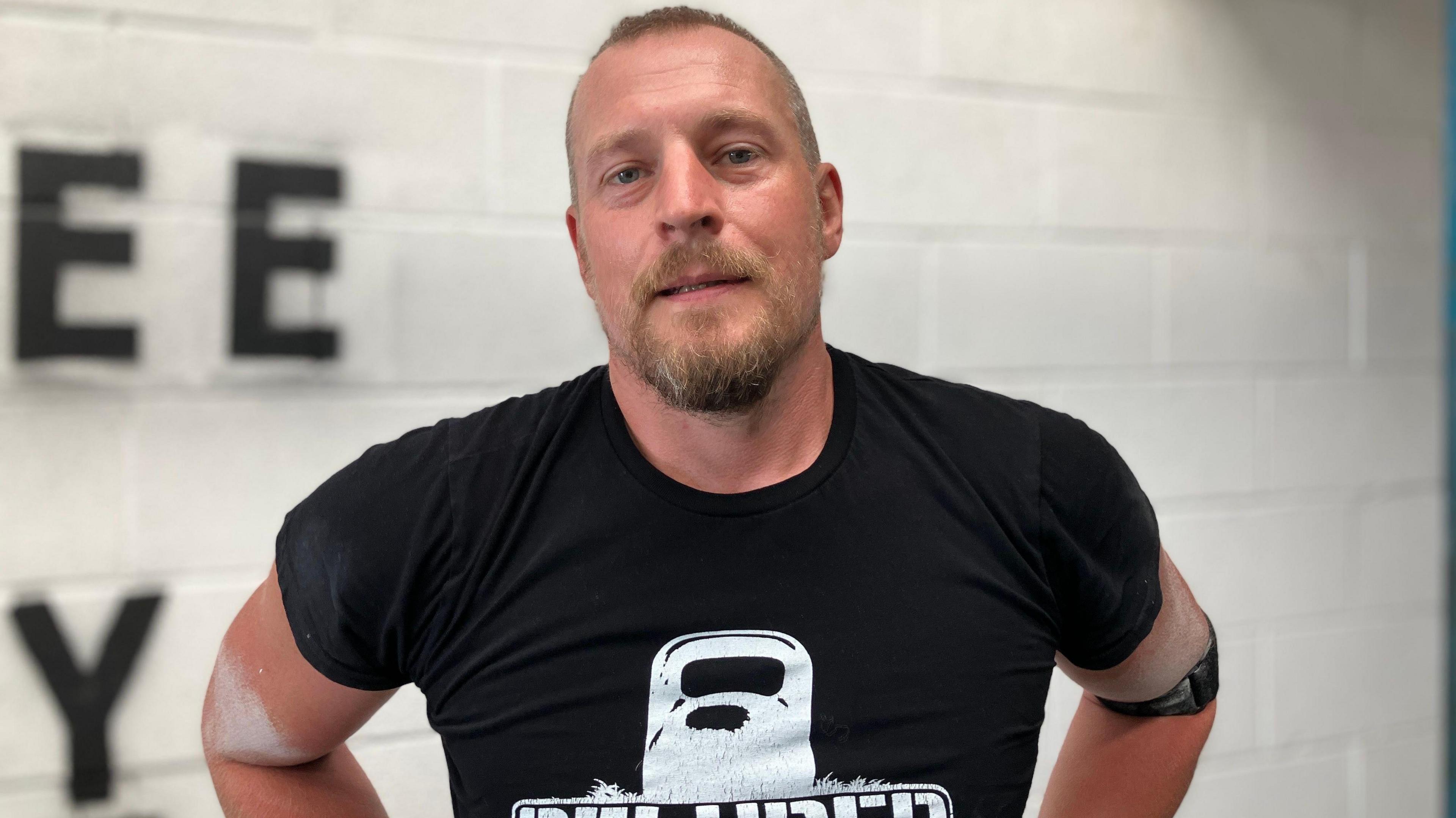 A man with short hair and a short beard and moustache is looking at directly at the camera. He wears a black t-shirt with an outline of a kettlebell on it