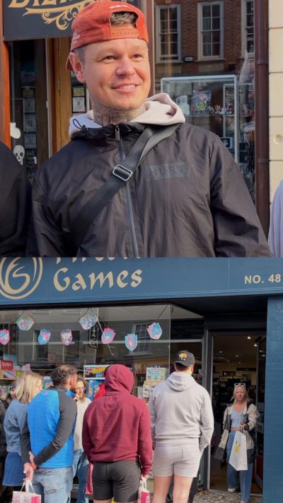 Man at the top of the image is smiling with a coat and backwards hat on. At the bottom of the image is people queueing at a game store. 