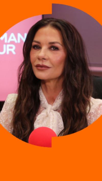 Catherine Zeta-Jones has long dark hair and is wearing a white top. She's sat in front of a red radio microphone.