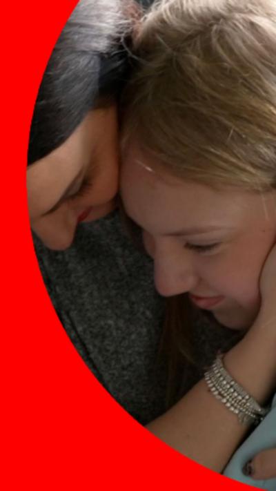 Two women are sat closely together with their heads touching. One woman rests a hand against the side of the other girls face, whilst they both look down.