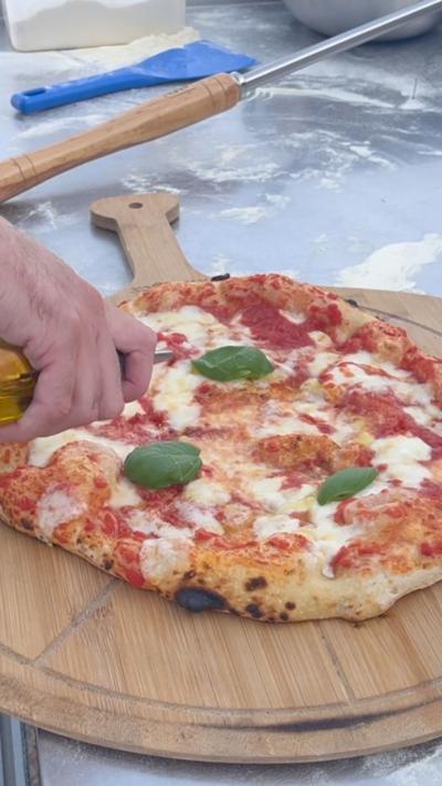 A tomato and cheese pizza on a wooden board. There is a man pouring olive oil on it. 