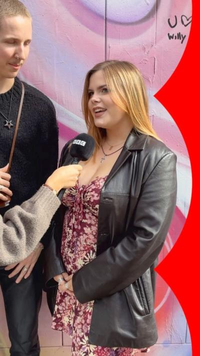A woman in a pink dress and black jacket speak to a microphone