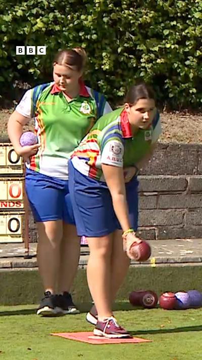 Two girls playing bowls with green polo shirts and blue shorts on.