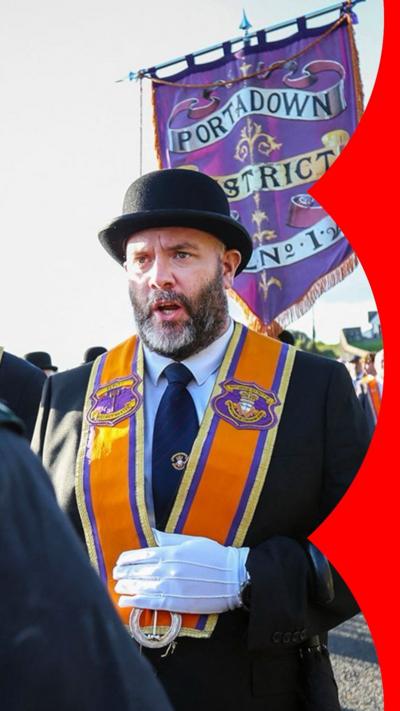 An Orange Order member talking to a police officer wearing a black suit, black hat and an orange sash. The rest of the lodge members are behind him with the church in the background. The police man is wearing a black and white checked hat with a black coat with green PSNI lapels.