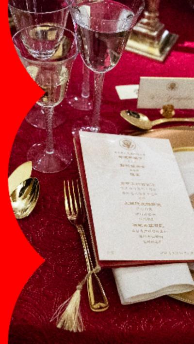 A dinner table on which there is a red table cloth, white and gold plates, gold cutlery, glasses of champaign and a white menu with gold writing.