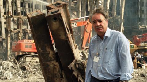 A man wearing a blue shirt and white ID badge stands in the destruction of the twin towers attack. There are metal girders sticking up and a digger behind them too.