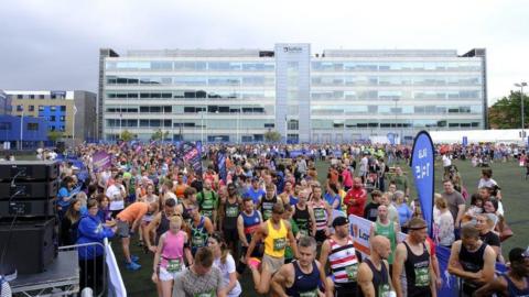Hundreds of runners gathered together ahead of the start of the Ipswich Half Marathon. A large glass building is in the background. 