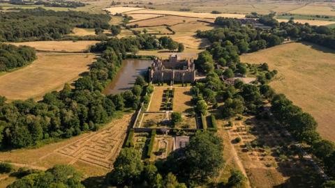 The Herstmonceux Castle estate. The castle can be seen from a bird's eye view. To the left of the castle is a river, and all around the castle are trees and shrubs. 
