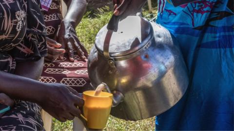 A close-up of someone's hand holding a large stove-top kettle and pouring a brown milky liquid into a yellow cup being held by someone else.