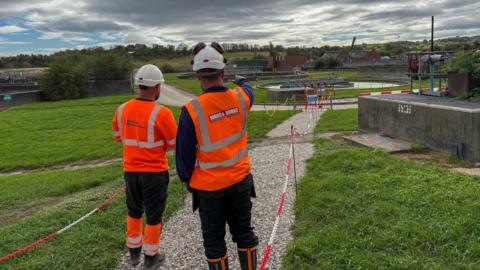 Two men in orange hi-vis jackets observe a waste treatment site from a footpath leading up to it. A small fence with red-and-white chains runs round the perimeter of the footpath.