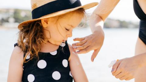 A woman in the right of the shot applies sunscreen to the arm of a little girl who wears a black and white spotted swimming costume and a straw hat with a black band.