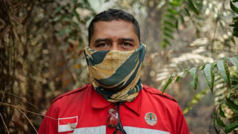 A man in red firefighting uniform patched with Indonesia's flag, and with a yellow and green neck gaiter pulled over his face, stands in a forest and looks into the camera