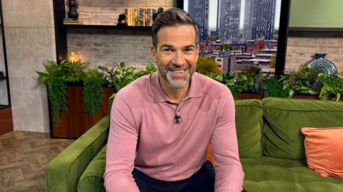 Gethin Jones stood in the Morning Live studio beside a TV displaying 'Morning Live'.