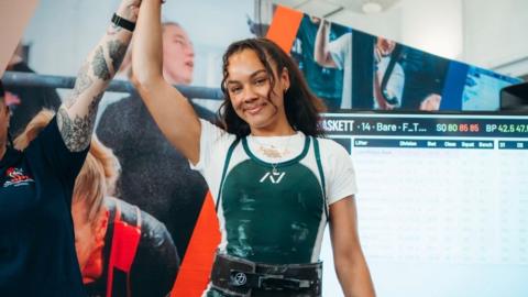 Ross is smiling wearing her weightlifting gear a mentor is lifting her arm up. 