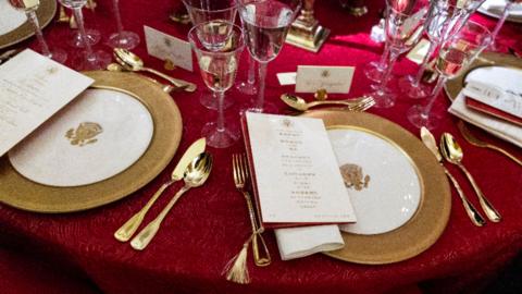 A dinner table on which there is a red table cloth, white and gold plates, gold cutlery, glasses of champaign and a white menu with gold writing.