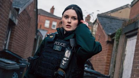 A young woman with brunette hair dressed in a dark green police uniform which includes a body-worn camera, stab vest and walkie-talkie. She's standing in an alleyway with red brick houses and bins alongside her. She's holding an ear piece to her left ear. She has a stunned impression on her face. 