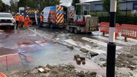 A burst water main on a zebra crossing on Lyde Road in Yeovil. There is water all over the road. Five men in orange high-vis coats and white hard hats are surveying the damage.