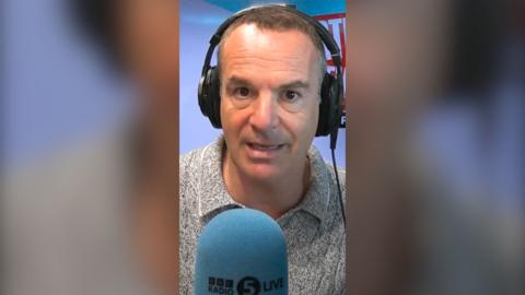 Martin Lewis looks into the camera. He is wearing headphones and is sat in front of a microphone with a blue mic muff. Only his face and shoulders are visible. 